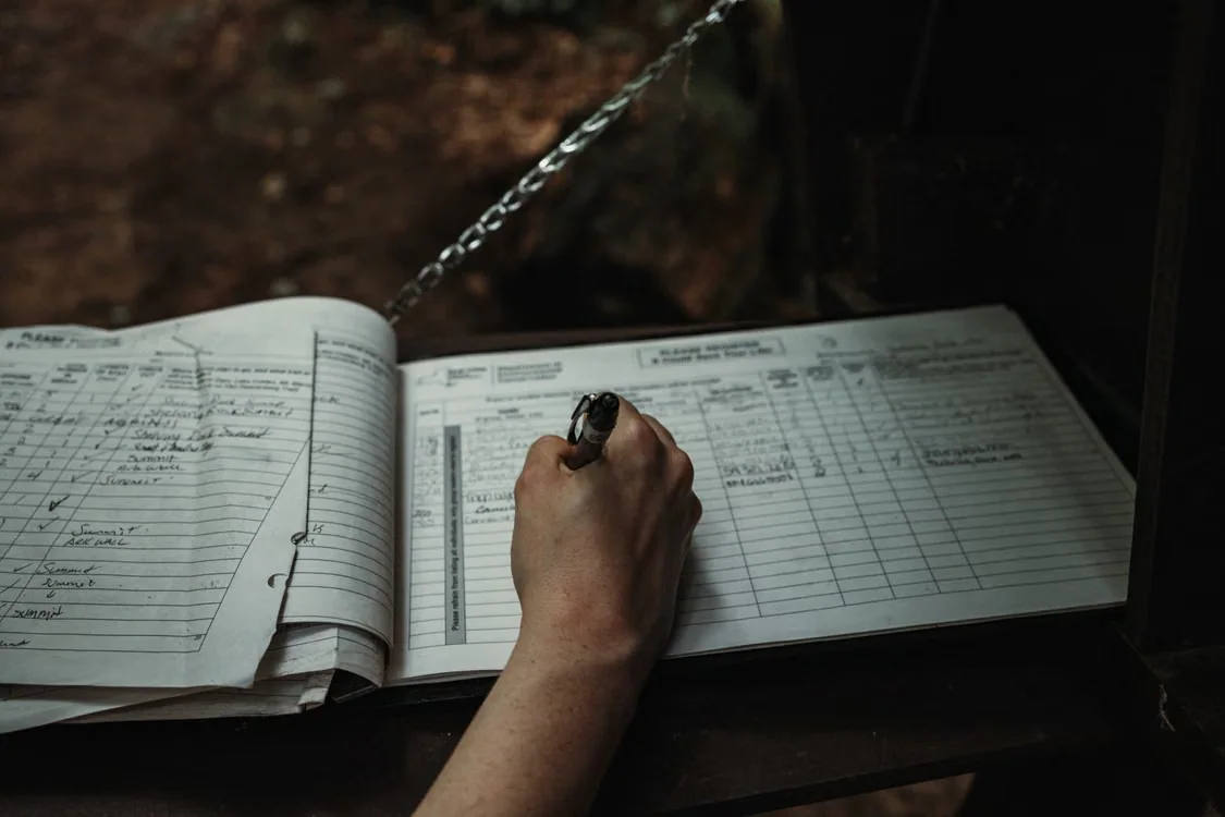 A hand writes notes in an open notebook with ruled, tabular pages.
